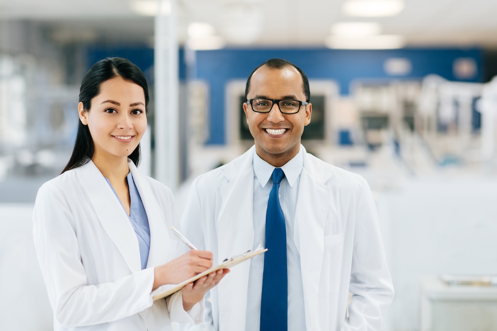 Two dentists in lab coasts smiling at the camera. One is writing on a clipboard