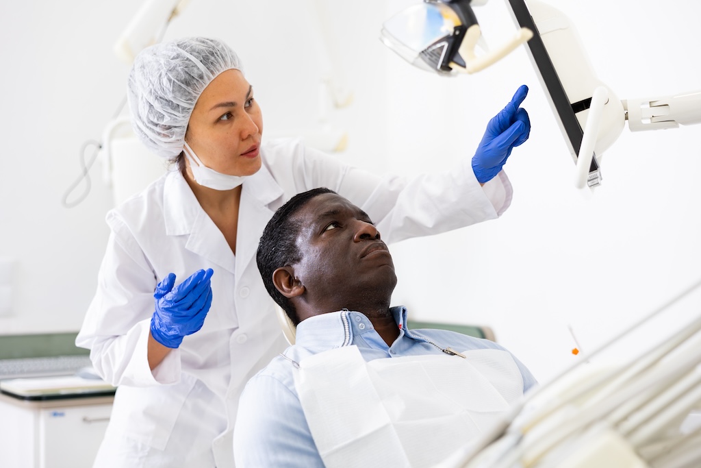 A dentist standing next to a patient who is sitting in a dental exam chair. The dentist is pointing to a screen on a monitor, showing the patient something on the screen