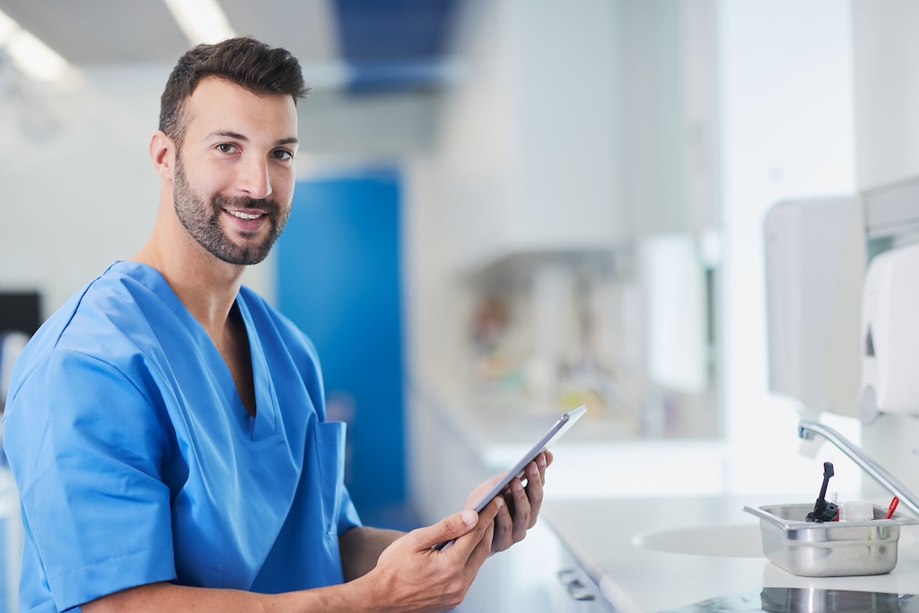 a dentist sitting in an exam room at a sink with a tablet in their hand. They are wearing scrubs and smiling at the camera